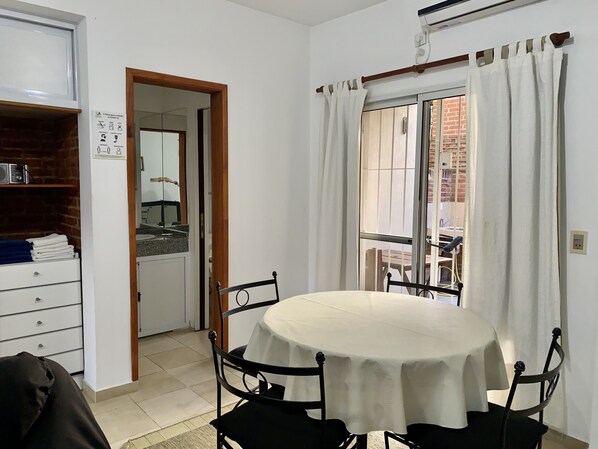 Un coin repas avec une table ronde recouverte d’une nappe blanche, entourée de quatre chaises. Il y a une fenêtre avec des rideaux blancs, une porte donnant sur une autre pièce et un climatiseur fixé au mur.