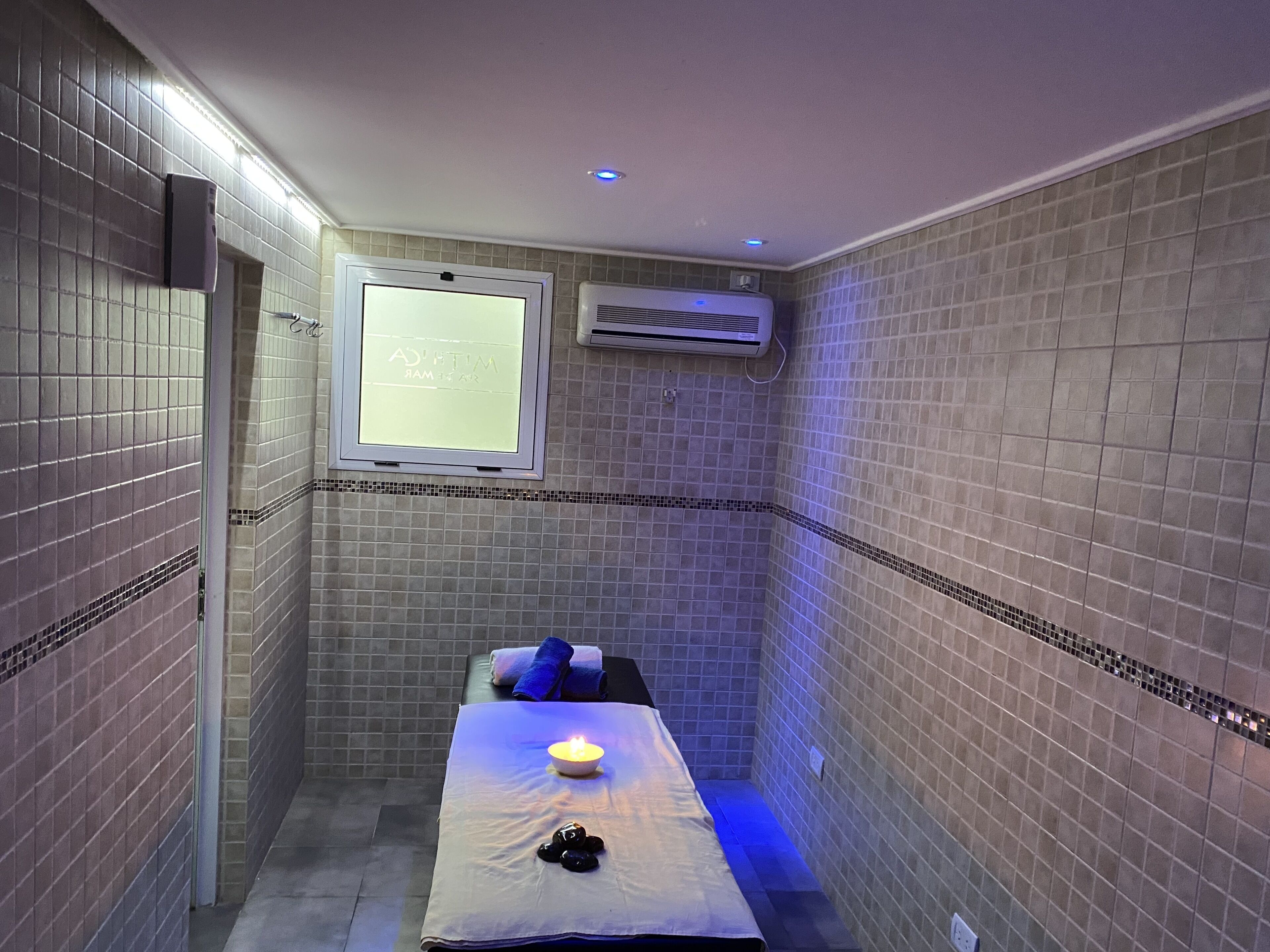Um pequeno quarto revestido de azulejos com uma mesa de massagem, uma vela acesa e uma janela com um letreiro.