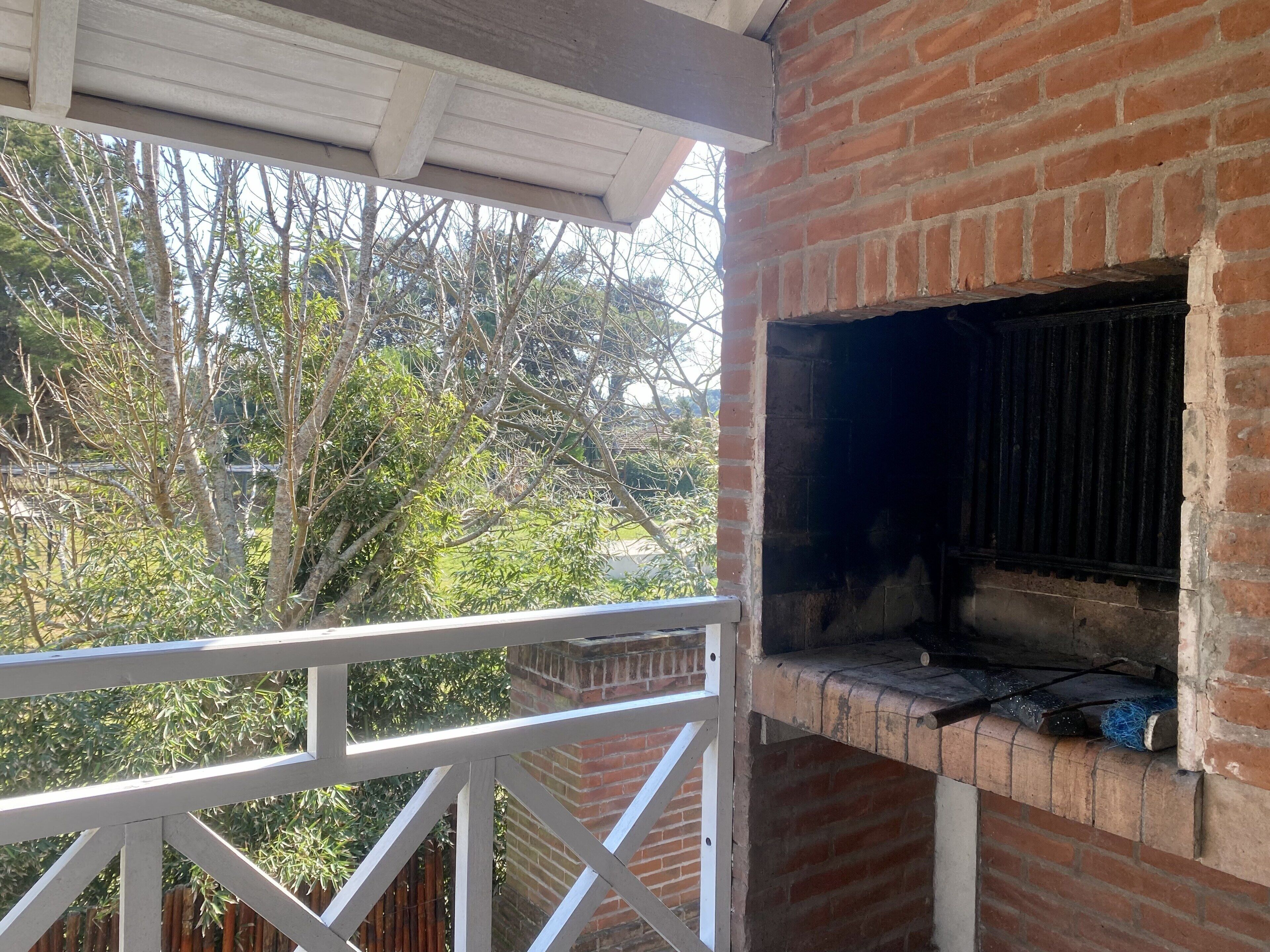 Área de churrasqueira ao ar livre com estrutura de tijolos, grade branca e vista para árvores e uma cerca ao fundo.