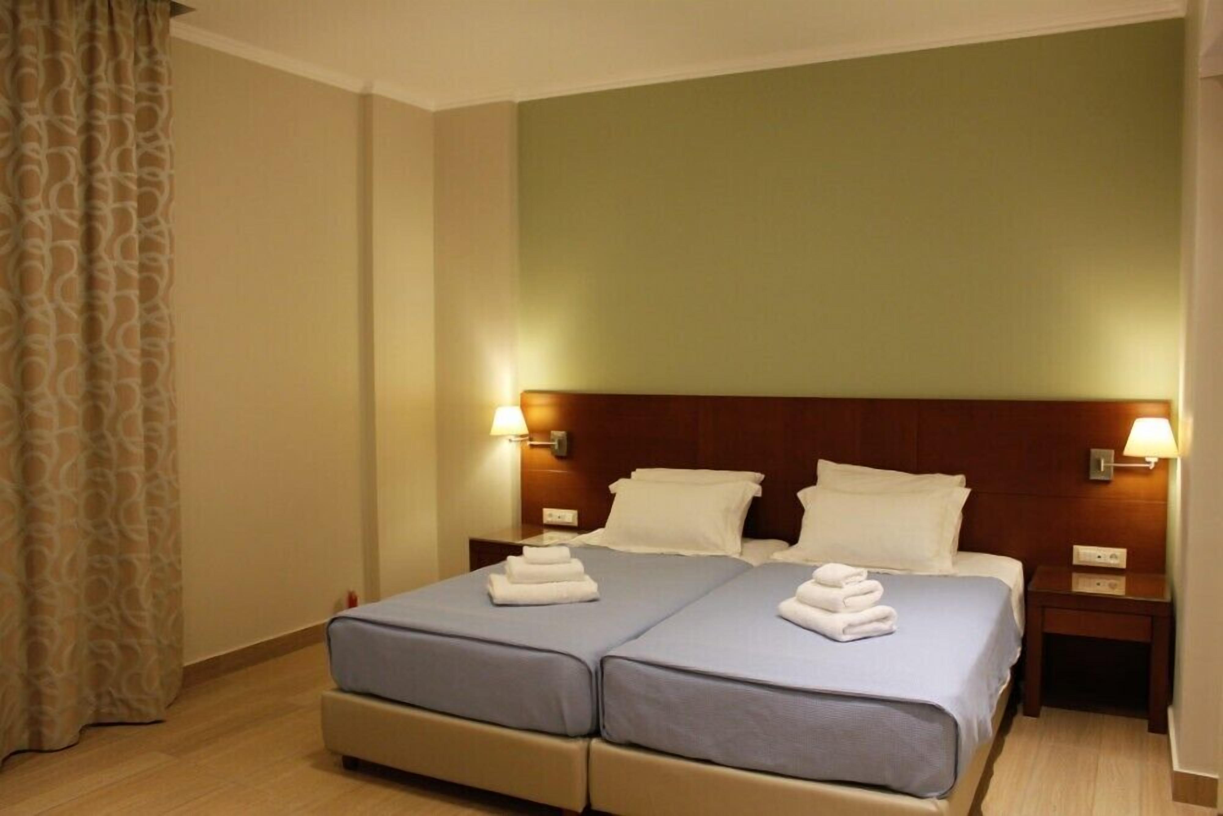 Une chambre double avec deux lits, chacun recouvert d'une couvre-lit bleue et agrémenté d'oreillers blancs, et dotée d'une tête de lit en bois. On y trouve deux tables de chevet avec des lampes, et un rideau est partiellement visible sur la gauche.