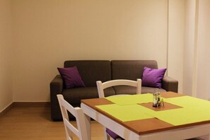 Ein kleiner, gemütlicher Raum mit einem Holztisch, zwei weißen Stühlen und einem braunen Sofa mit violetten Kissen.