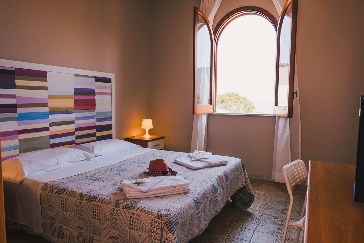 A hotel room with a bed, a nightstand with a lamp, a chair, and a window with curtains.
