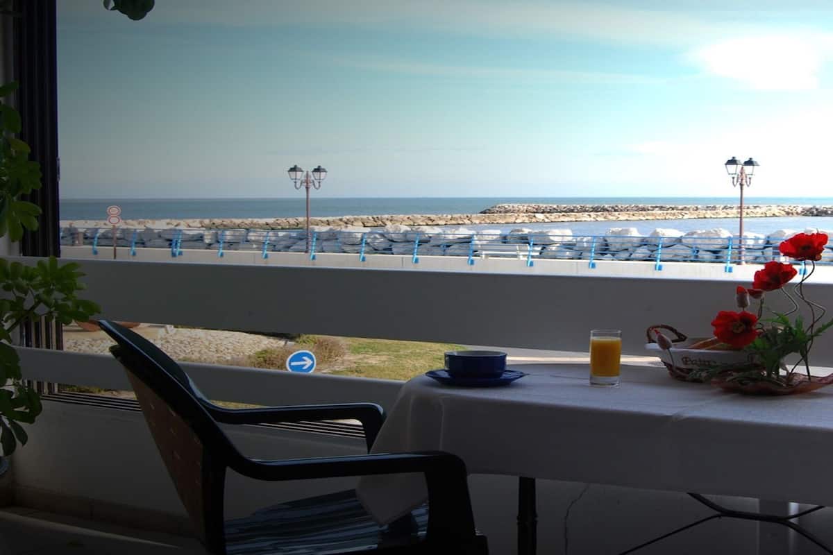 A table with a view of the beach, a chair, a tablecloth, a glass of orange juice, and a vase with red flowers.