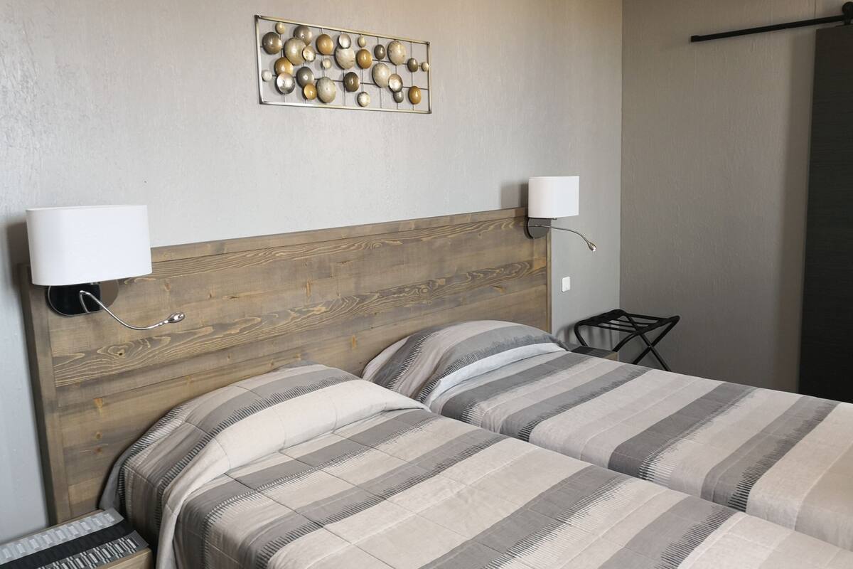 A bedroom with two beds, wooden headboard, wall-mounted lamps, and a decorative wall piece.