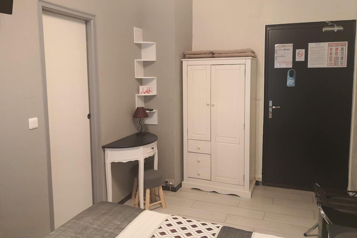 A small room with a desk, chair, and wardrobe. There is a door with a lock and a small shelf unit against the wall.