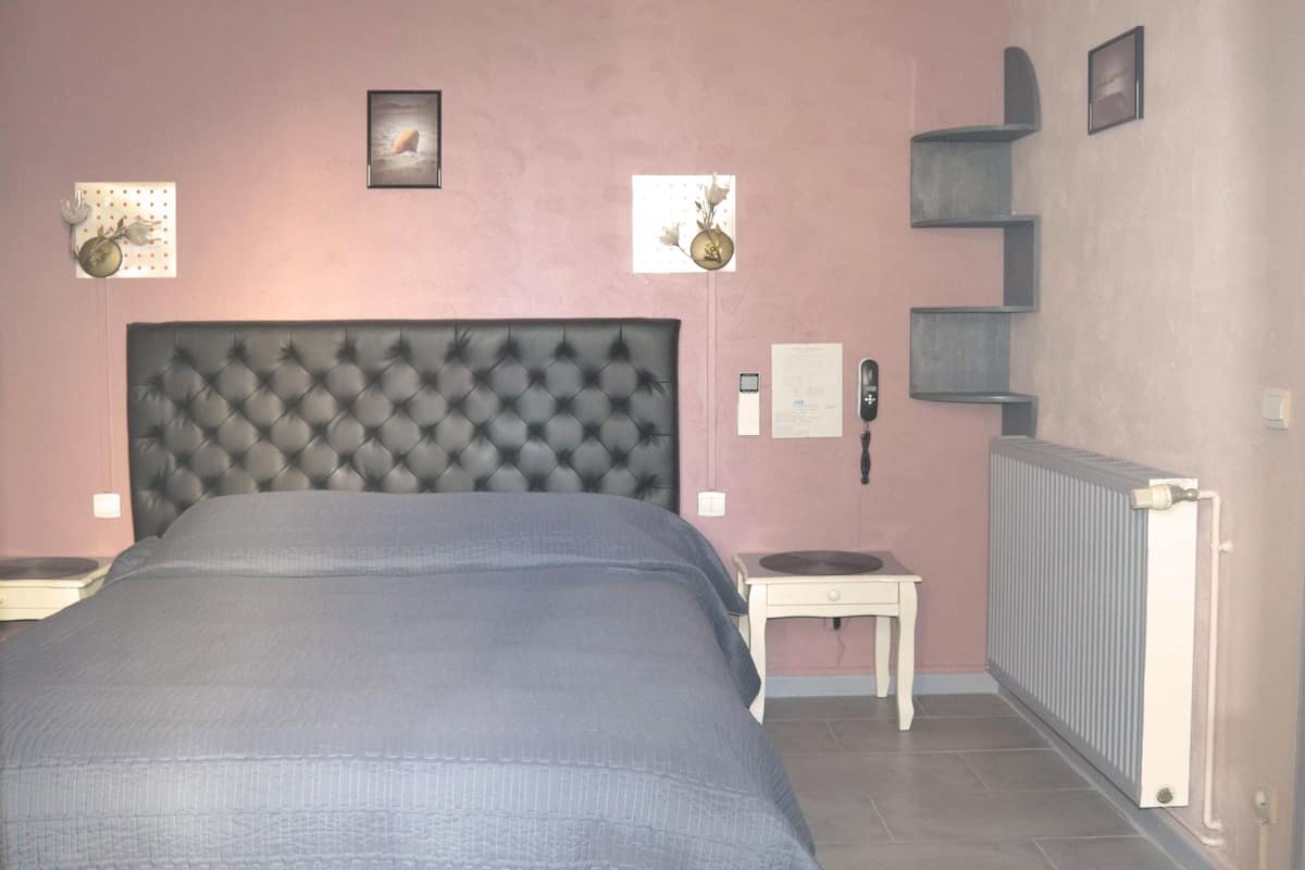 A bedroom with a tufted headboard, a bedside table, a radiator, and wall-mounted shelves.