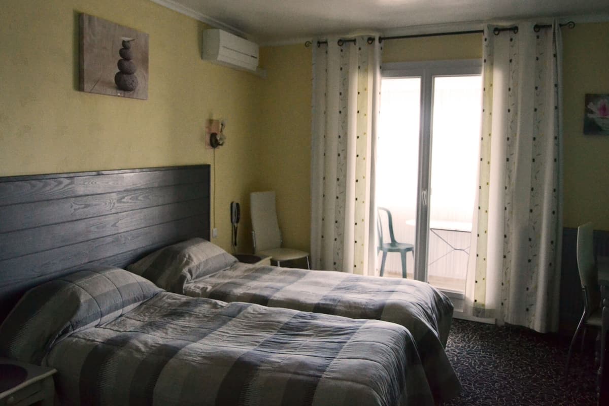 A hotel room with two beds, a chair, a table, and a view of the outside through a window with curtains.