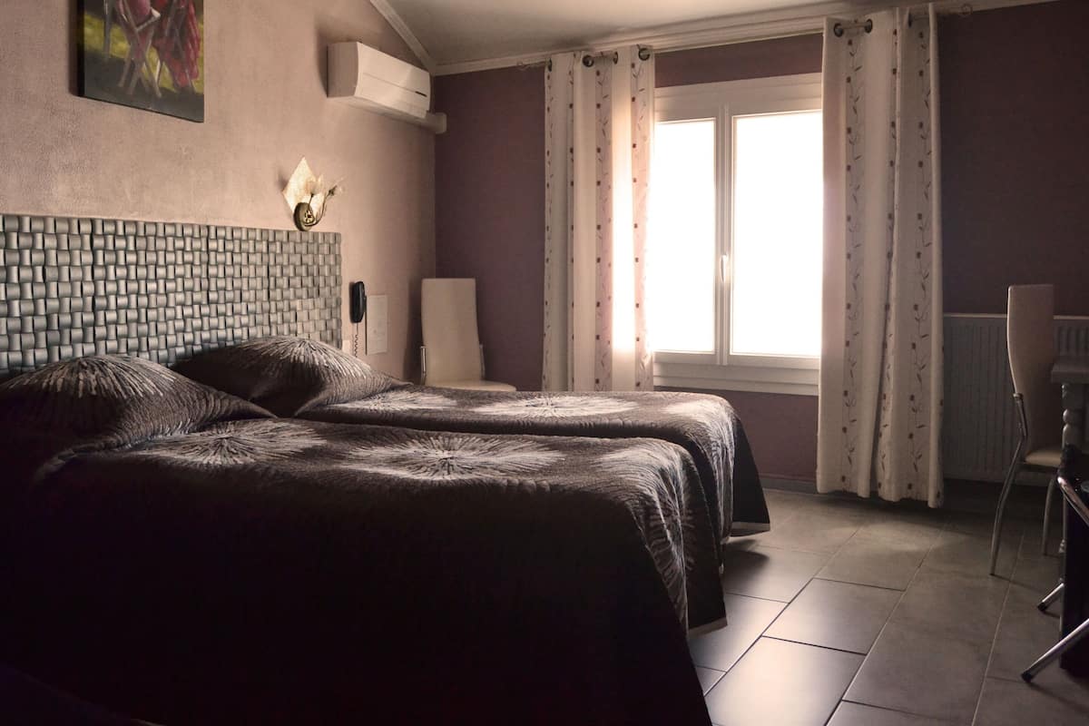 A hotel room with two beds, a chair, a desk, a window with curtains, and a wall-mounted phone.