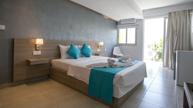 A modern hotel room with a large bed, a nightstand, a lamp, a window with curtains, and a view of greenery outside. - Flora Maria Hotel (Ayia Napa)