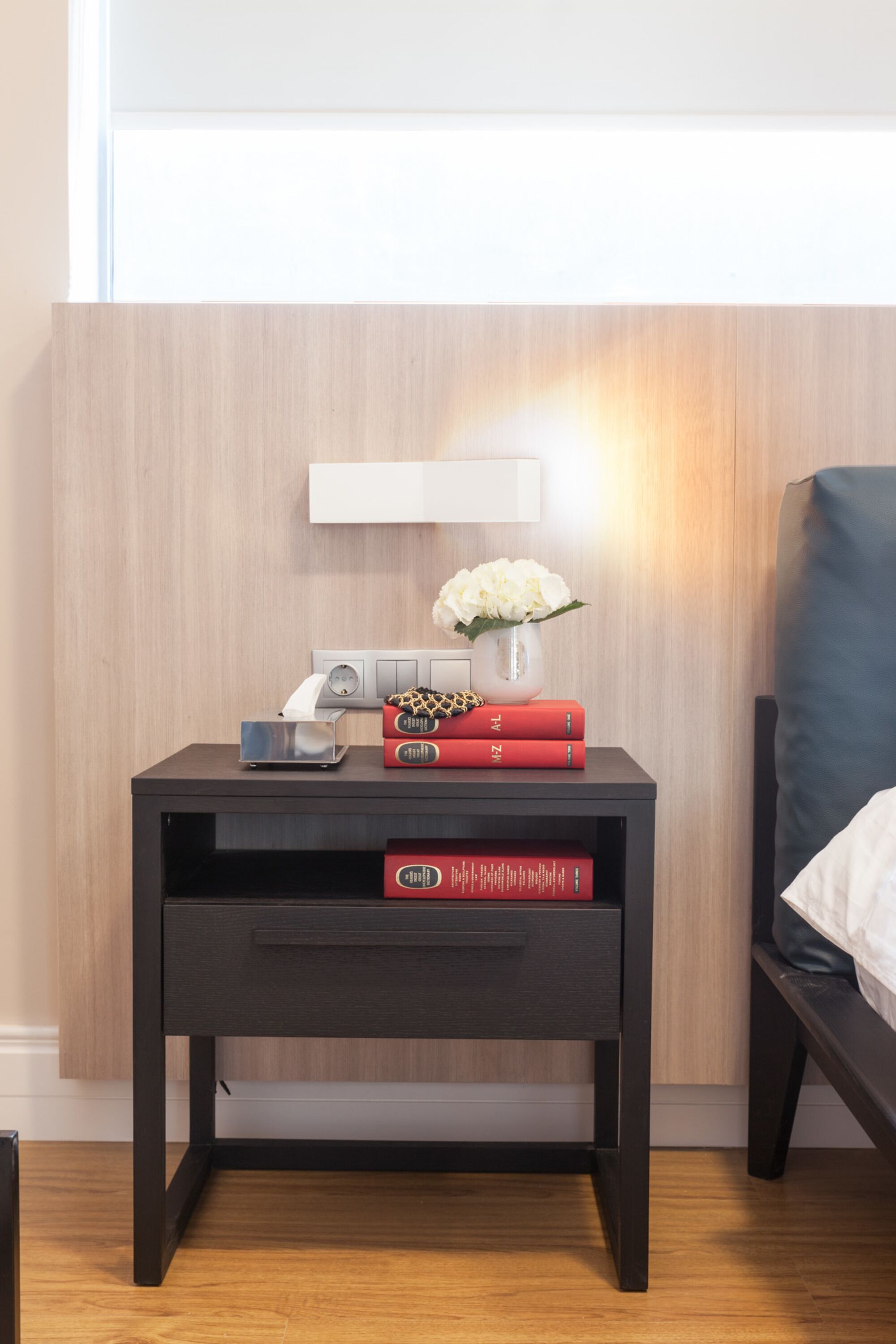 A modern bedroom with a wooden floor, a dark nightstand with a lamp, a vase with flowers, and a bed with a blue headboard.