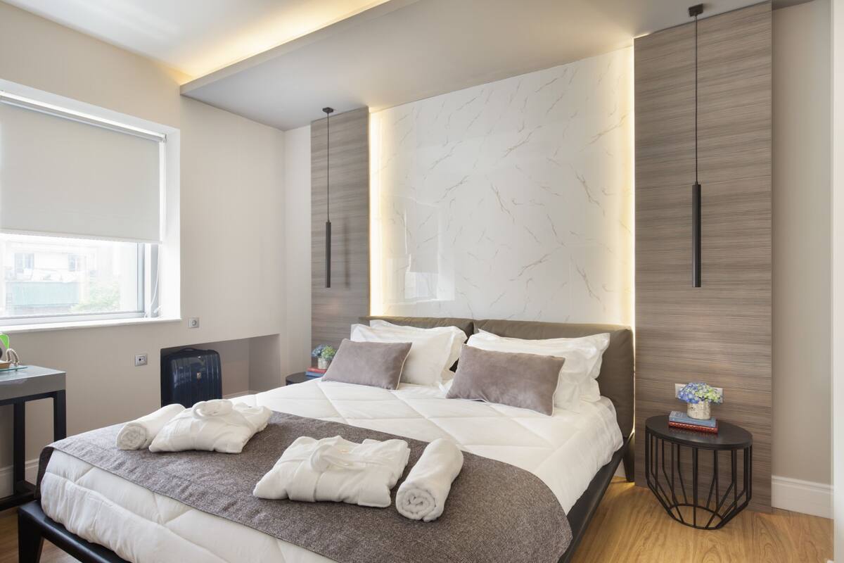 Ein modernes Hotelzimmer mit einem großen Bett, einem Nachttisch, einem kleinen Seitentisch und einem Fenster mit Blick.