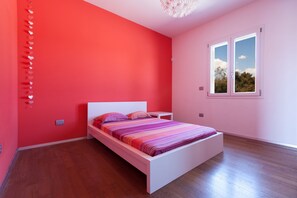 Une chambre avec un lit blanc, des murs roses et une fenêtre donnant sur des arbres.