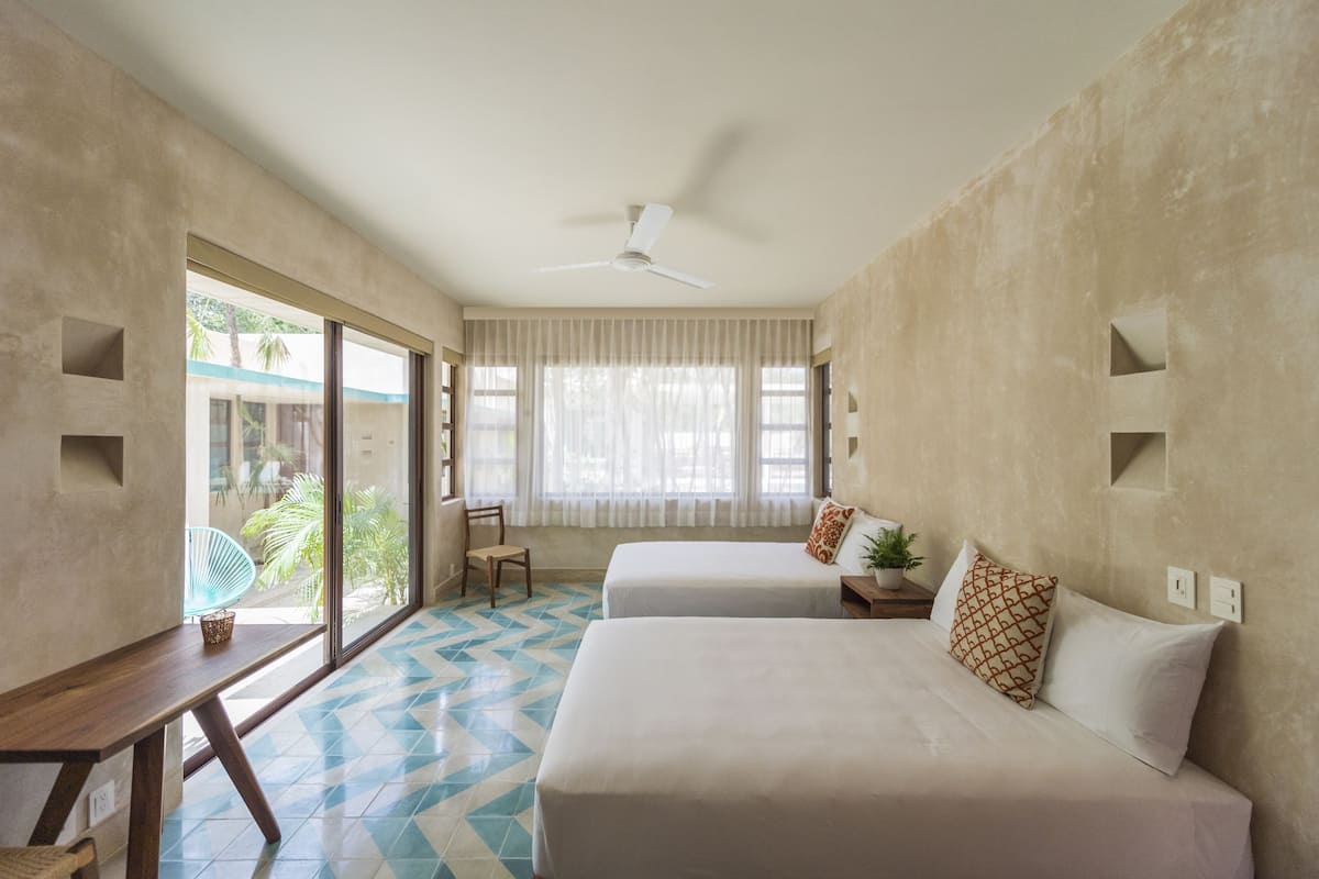 Une chambre d’hôtel avec deux lits, un ventilateur de plafond, une fenêtre avec des rideaux, une chaise et une petite table avec une plante.