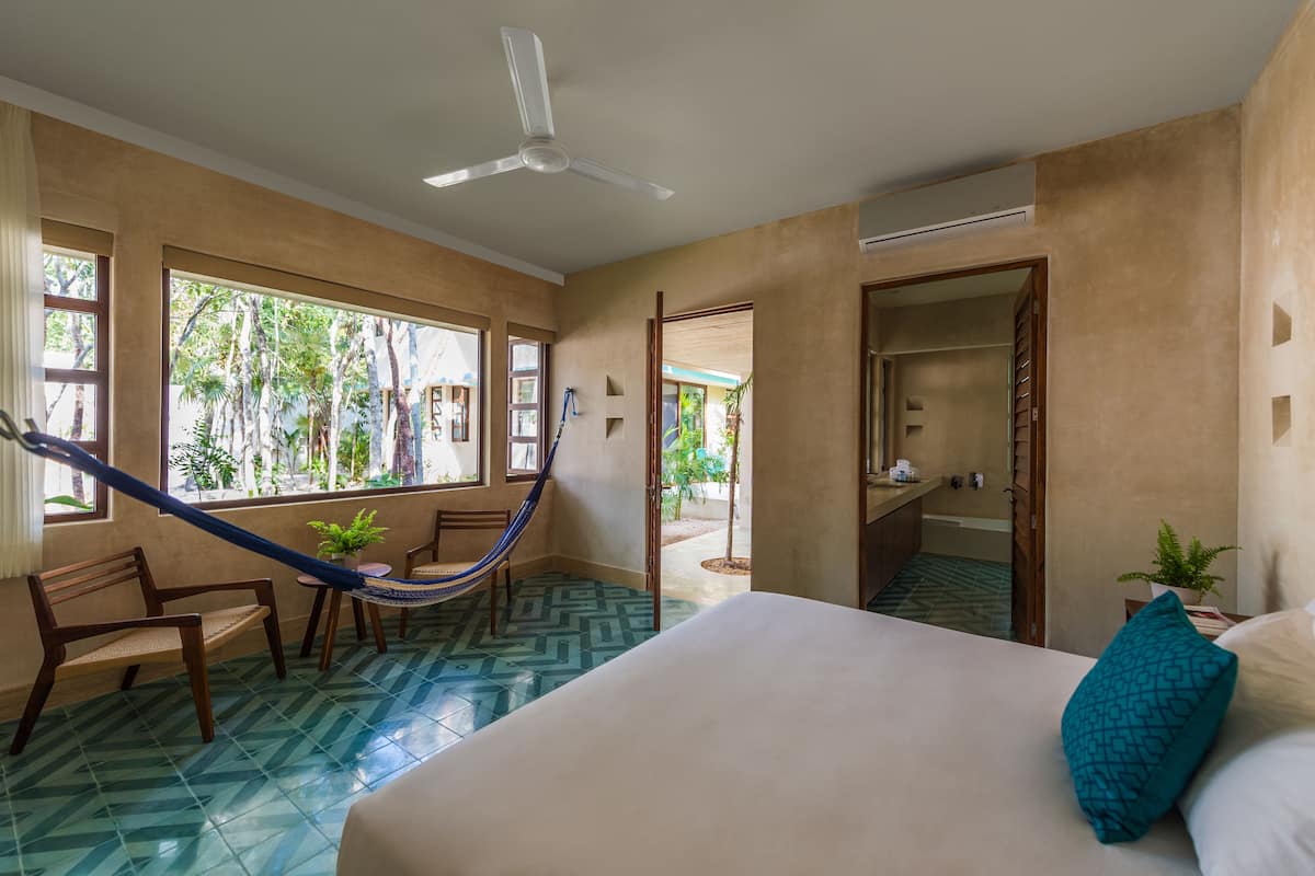 Une chambre d’hôtel avec un lit, un hamac, un ventilateur de plafond, une grande fenêtre et une vue sur l’extérieur.