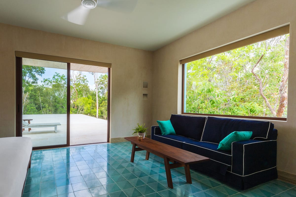 Une pièce moderne dotée d’une grande porte-fenêtre coulissante donnant sur un espace extérieur, d’un canapé bleu foncé agrémenté de coussins turquoise, d’un banc en bois et d’une petite table sur laquelle est posée une plante.