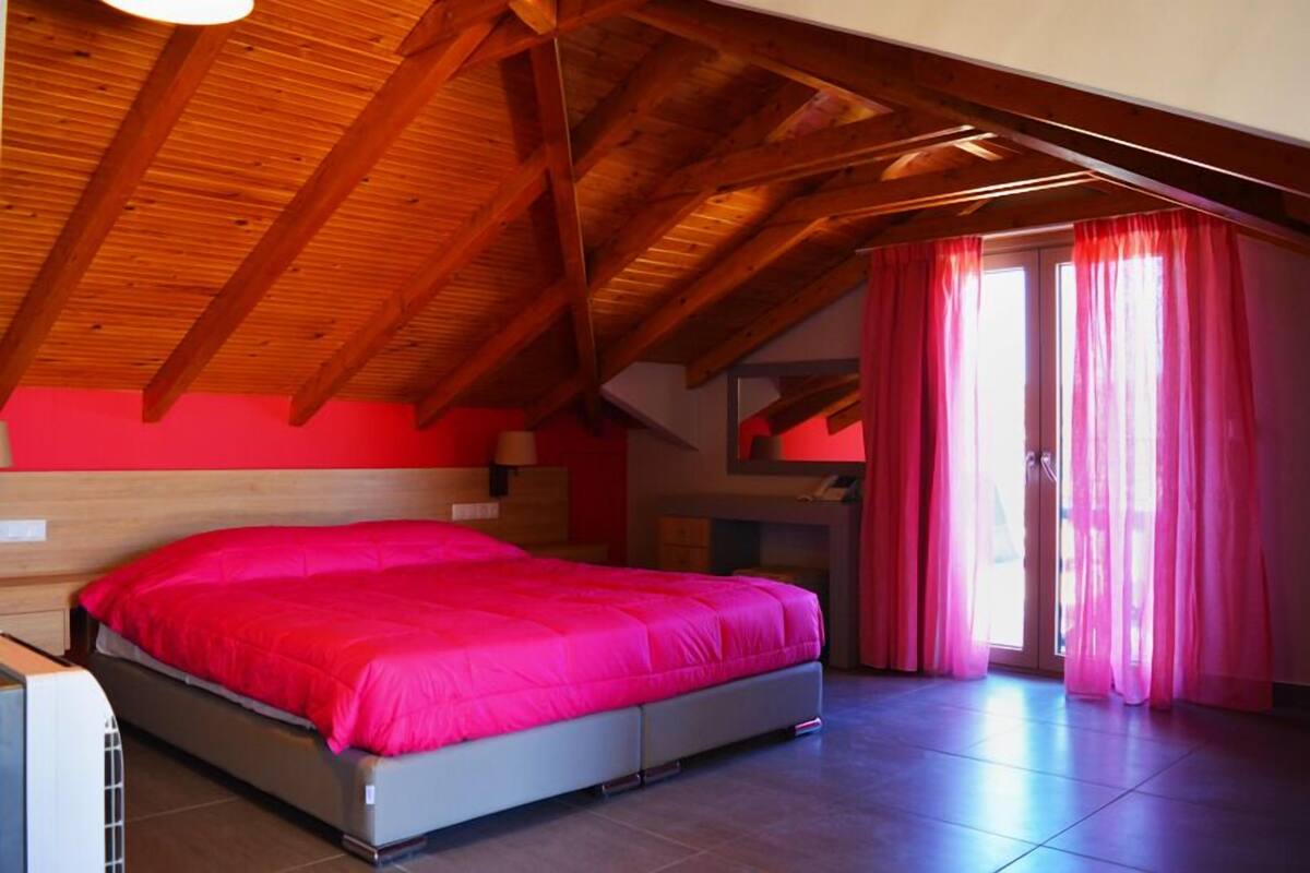 A bedroom with a wooden ceiling, a bed with a pink bedspread, and a sliding glass door with pink curtains.