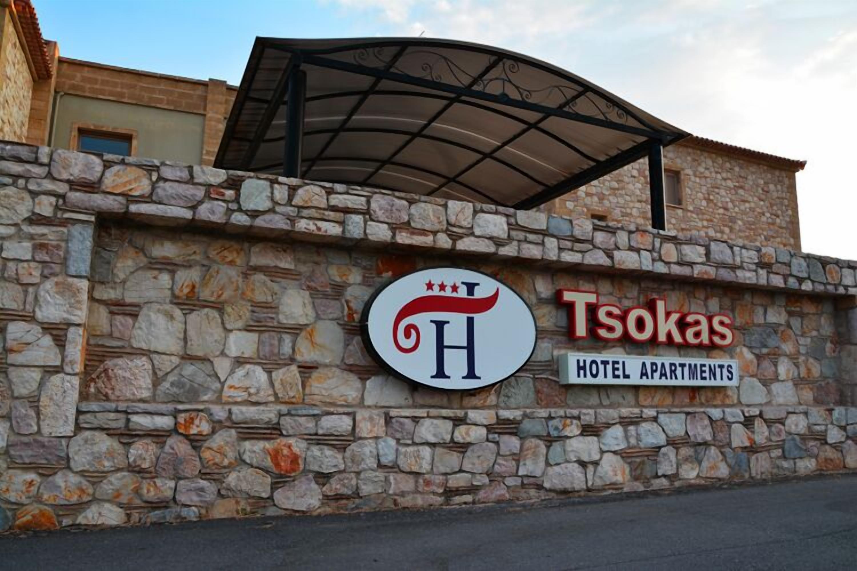 A stone wall with a sign for Tsokas Hotel Apartments, featuring a crown logo and a curved roof structure above.