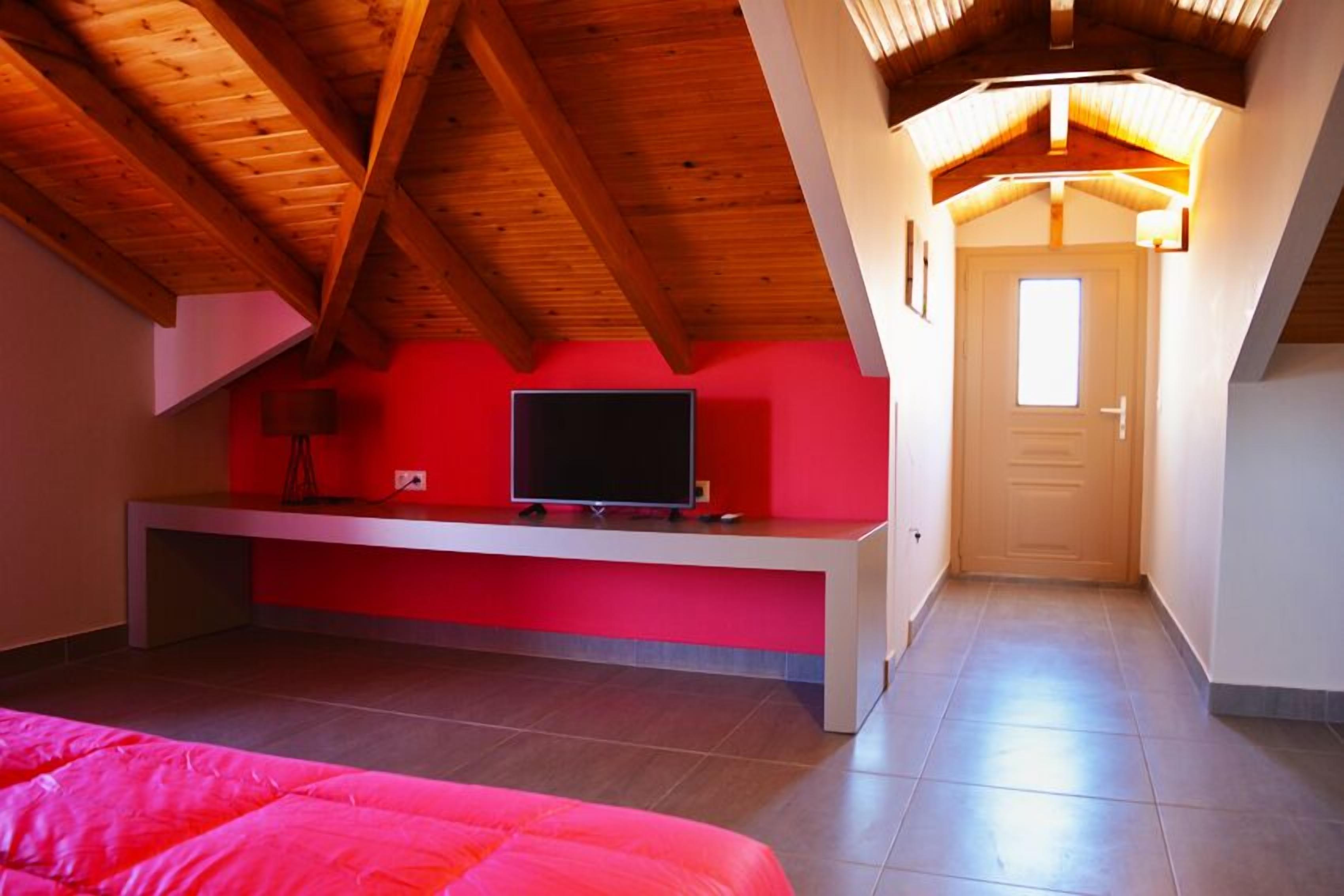 A bedroom with a sloped ceiling, a bed, a TV on a stand, and a door leading to another room.