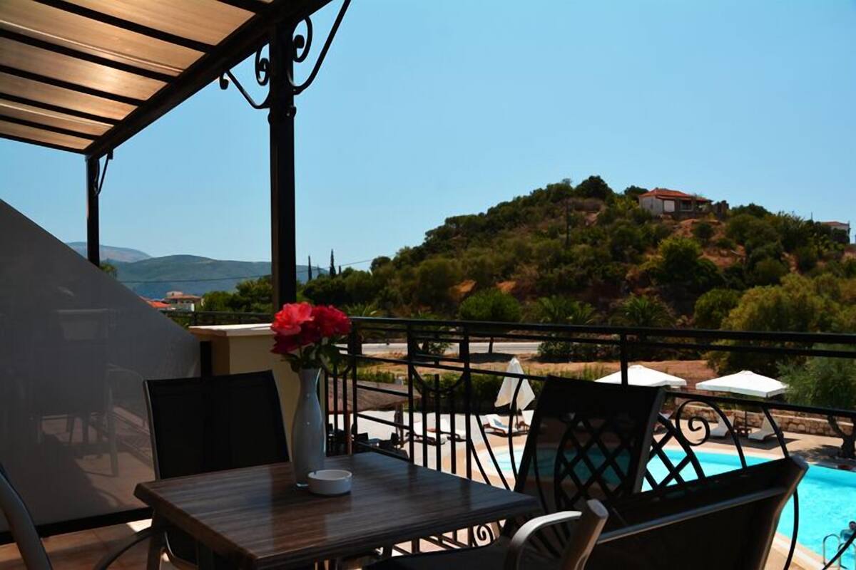 A terrace with a table, chairs, a vase with flowers, and a view of a swimming pool and hillside.