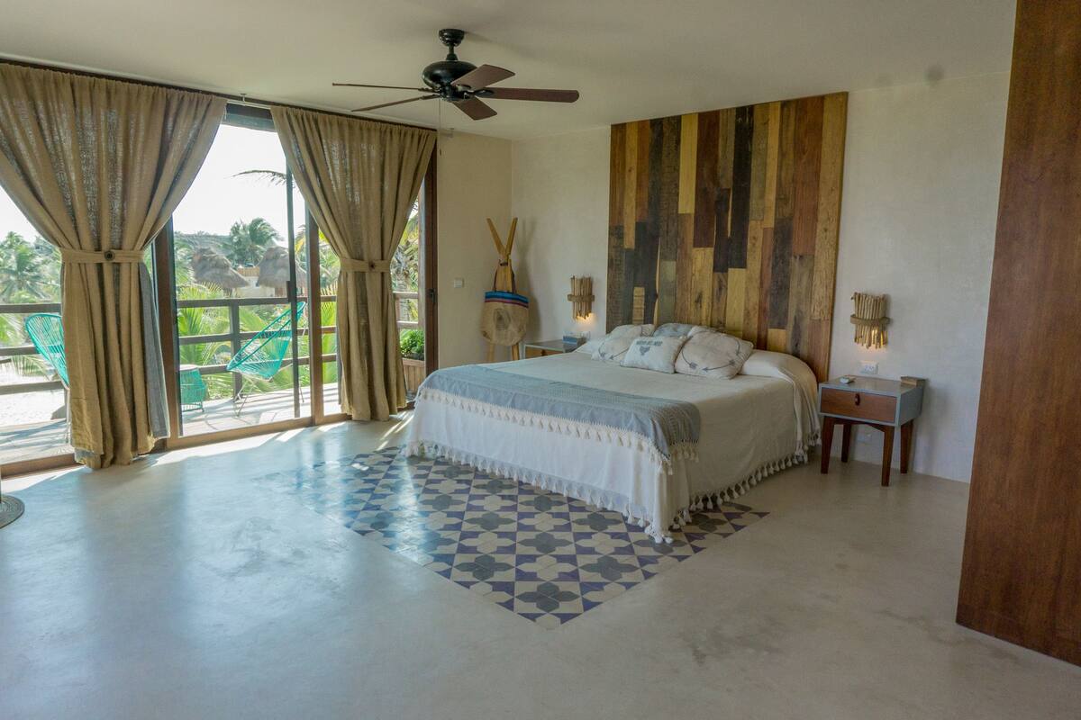 A bedroom with a bed, a ceiling fan, a wooden wall panel, a nightstand, and a window with curtains.