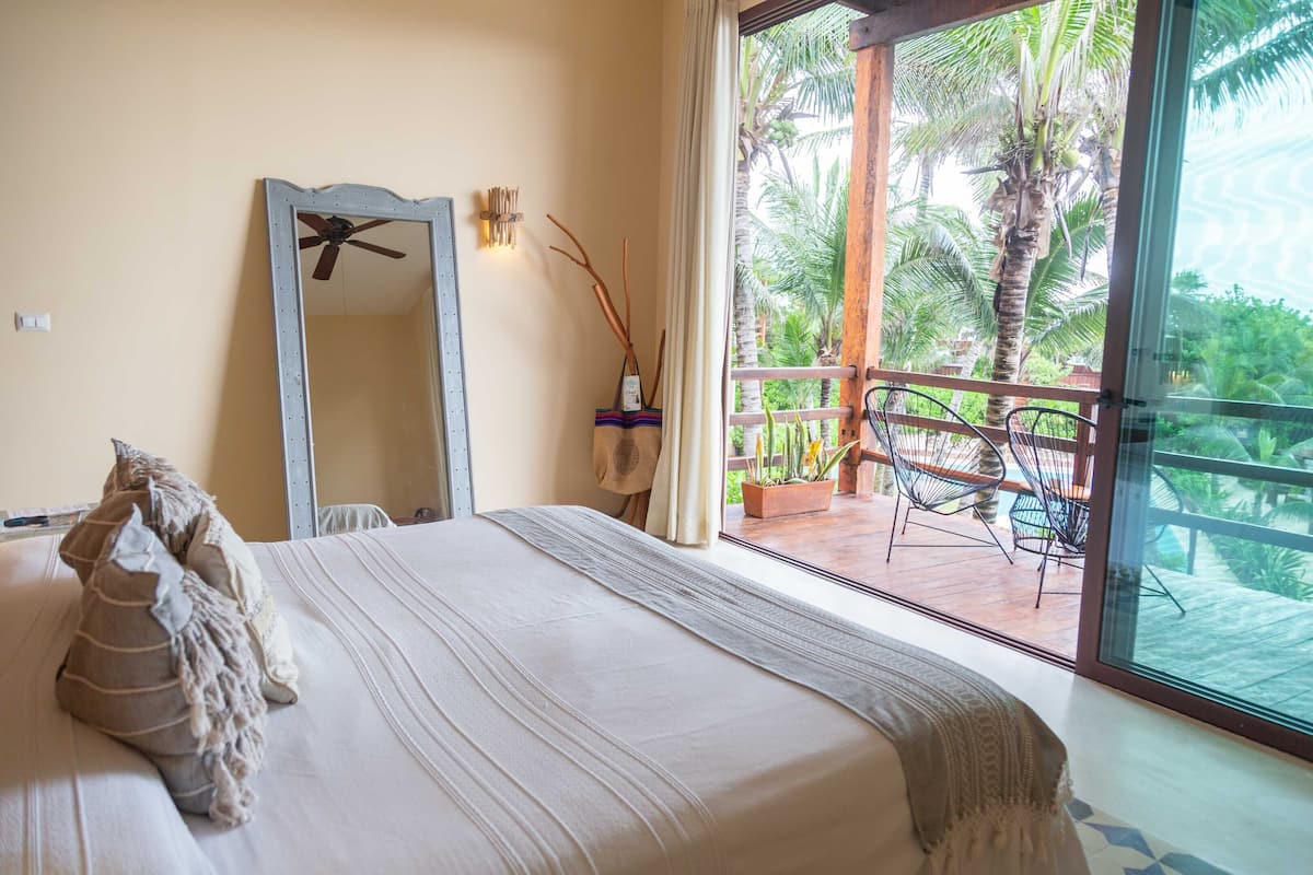 A bedroom with a large bed, a ceiling fan, a mirror, and a view of a balcony with chairs and palm trees.
