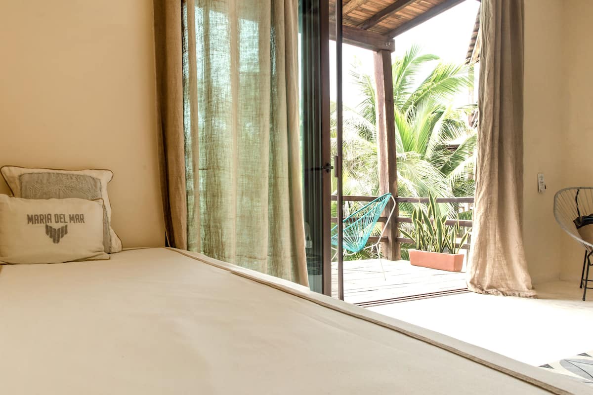 A room with a bed, a window with curtains, a chair, and a view of palm trees.