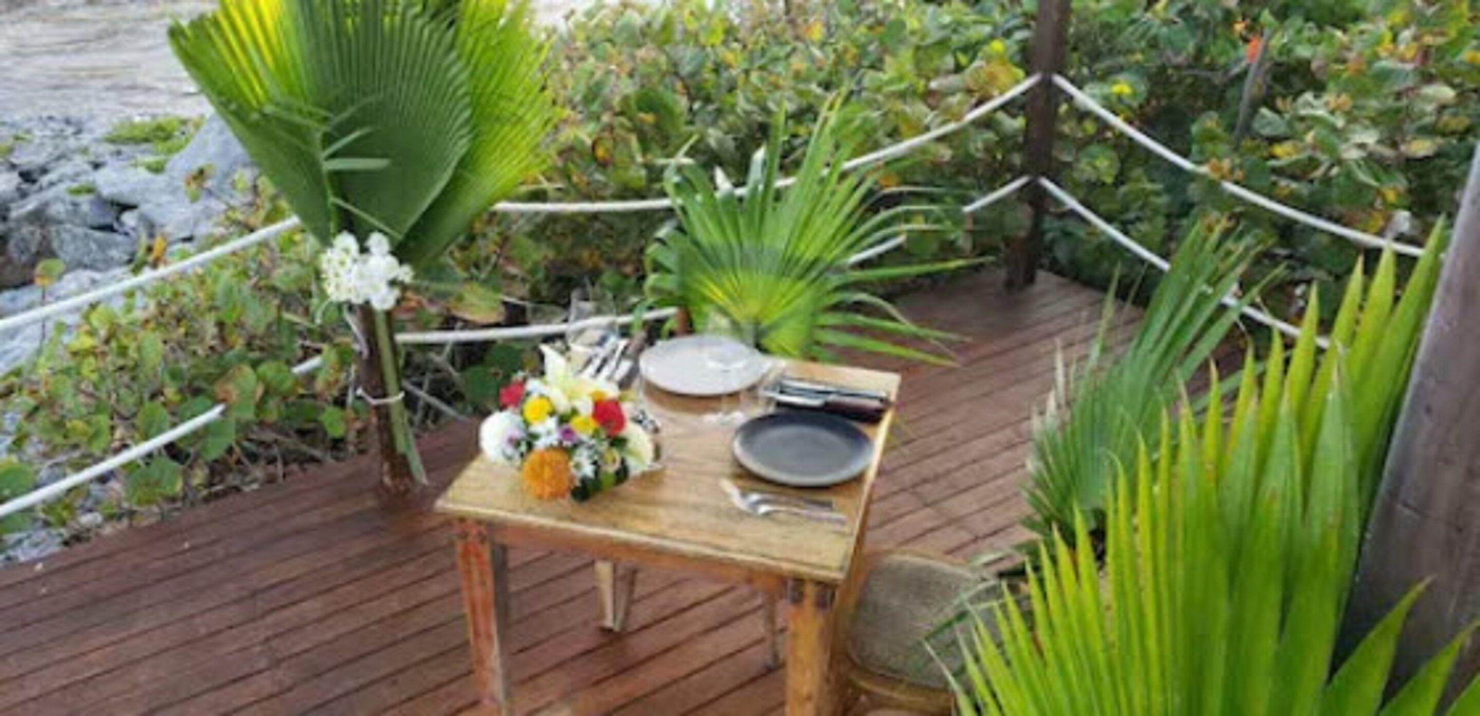 A wooden table set outdoors with a floral arrangement, a plate, and cutlery.