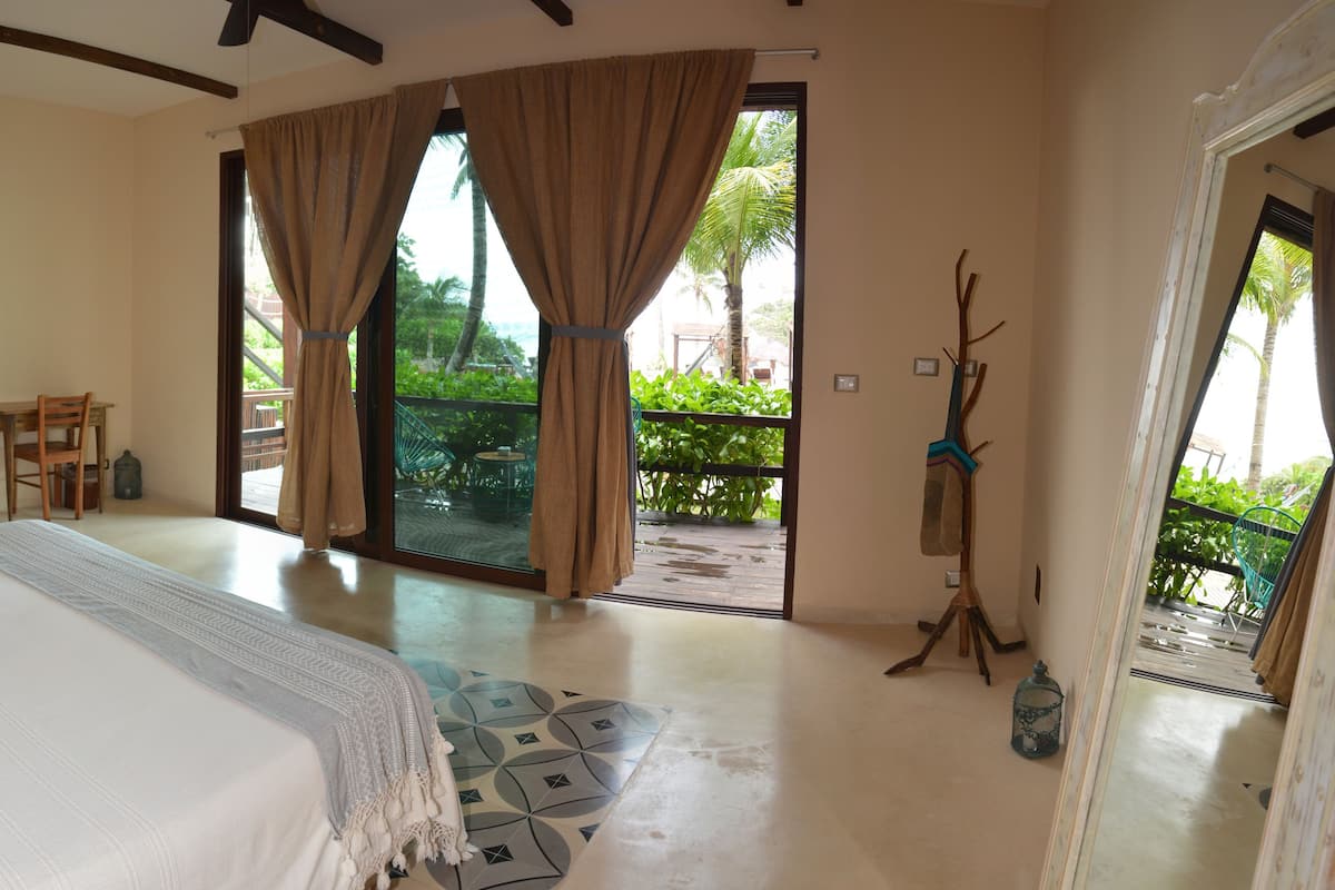 A spacious bedroom with a large bed, a chair, and a table. There is a balcony with a view of greenery.