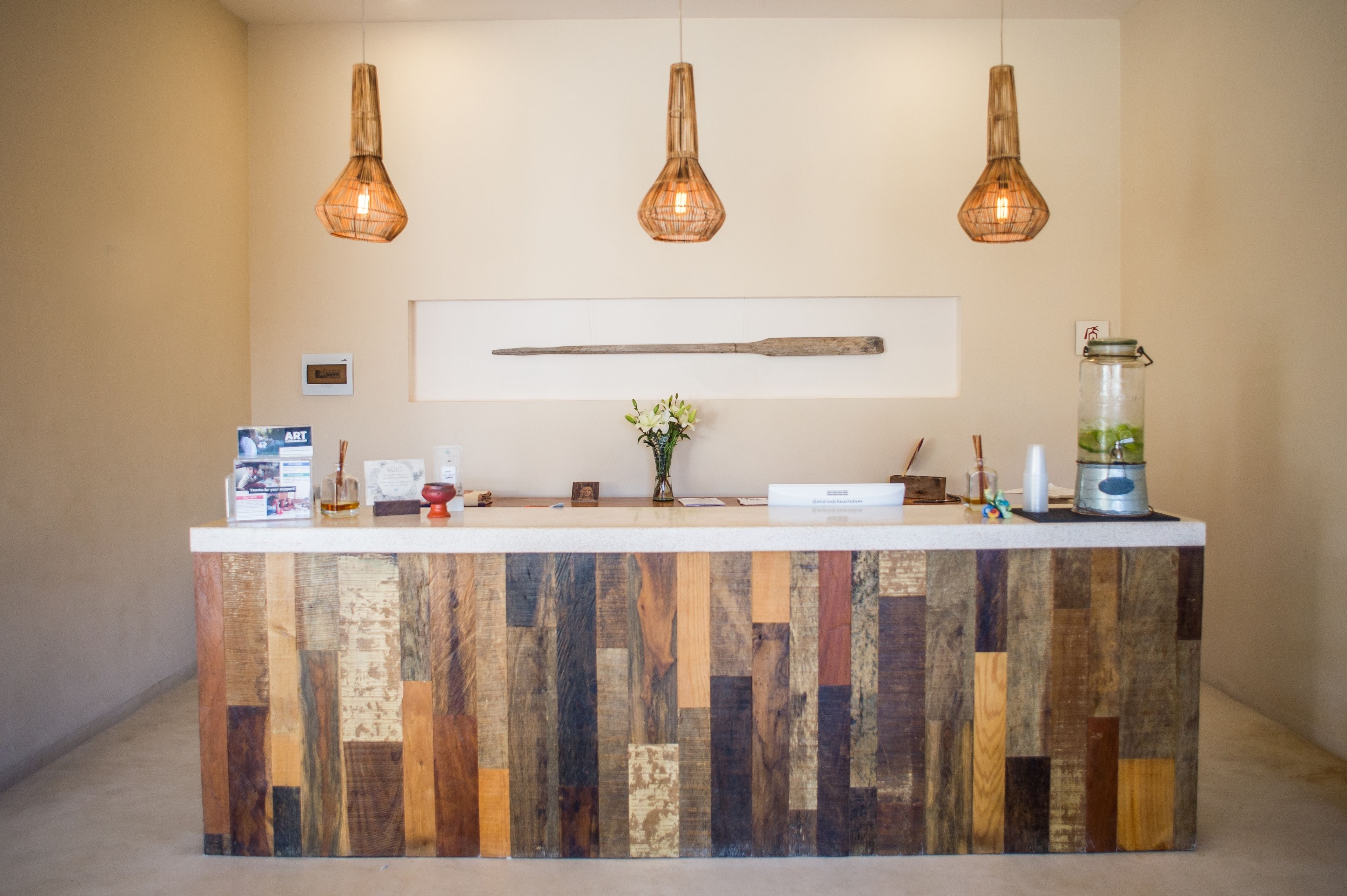 Un mostrador de recepción con diseño de tablones de madera, tres lámparas colgantes, una obra de arte enmarcada y un pequeño jarrón con flores.