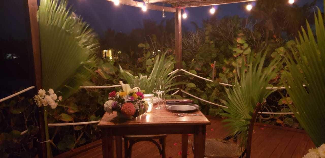 Un espace de restauration en plein air avec une table en bois dressée pour deux personnes, entouré d’une végétation luxuriante et illuminé de guirlandes lumineuses.