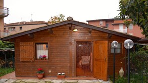 Una cabaña de madera con techo inclinado, puerta principal, ventana con contraventanas y un pequeño porche con plantas en macetas.