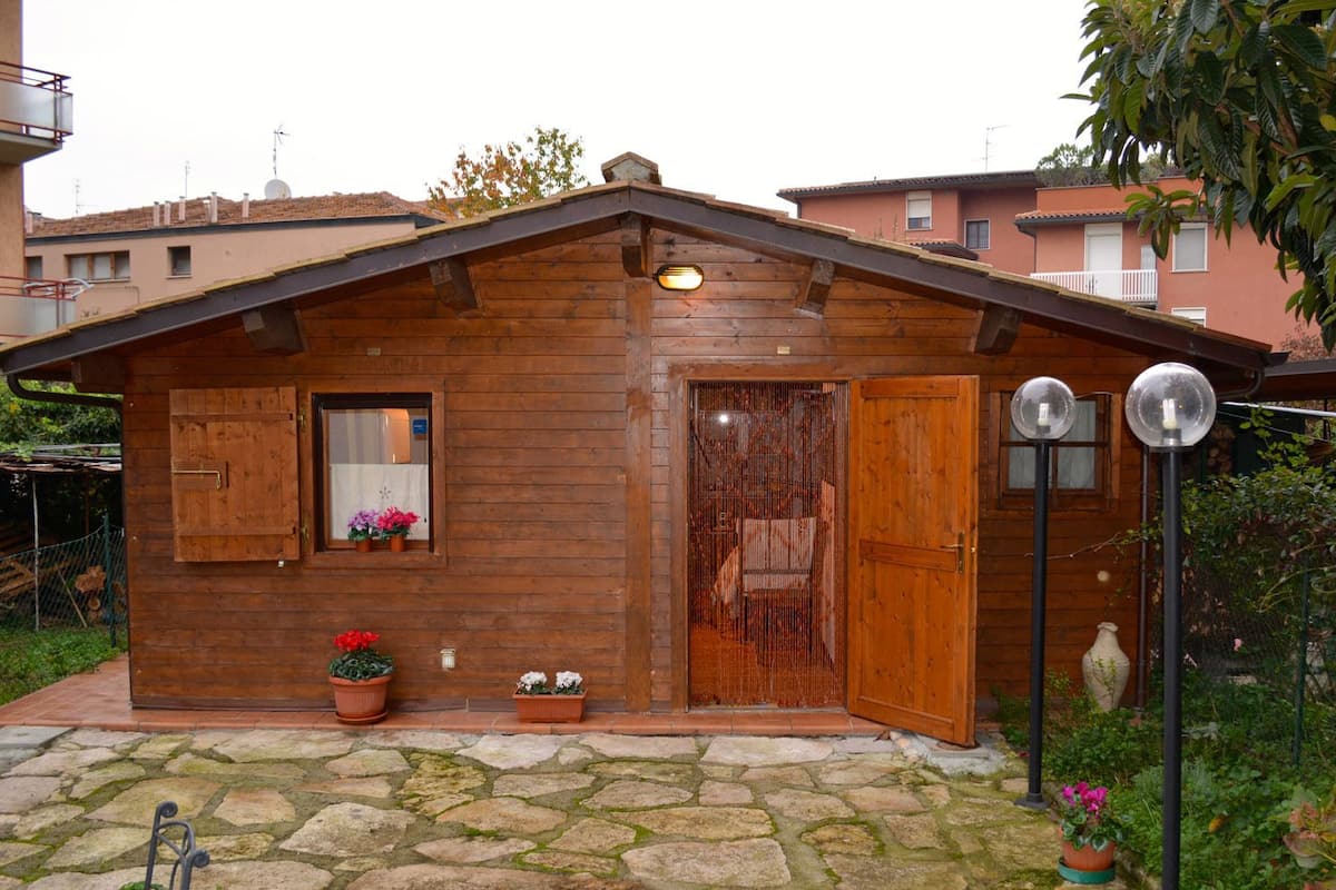A wooden cabin with a sloped roof, a front door, a window with shutters, and a small porch area with potted plants.