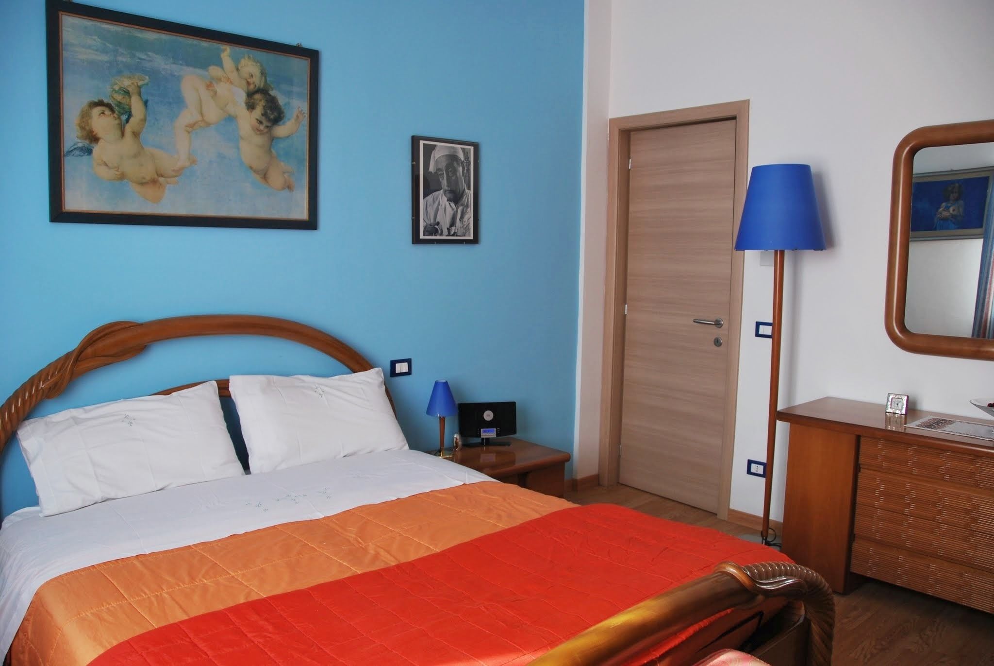 A hotel room with a bed, a nightstand, a lamp, a dresser, a door, and two framed pictures on the wall.