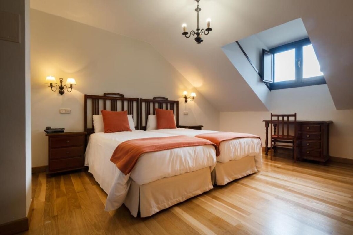 A bedroom with a sloped ceiling, a bed with two pillows, a nightstand, a dresser, and a desk.