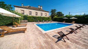 A swimming pool with wooden deck chairs and umbrellas, surrounded by greenery and a stone building in the background. - La Posada de Pradorey (Gumiel de Mercado)