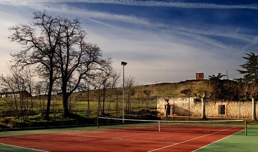 Una cancha de tenis con una red, rodeada de árboles y un muro de piedra.