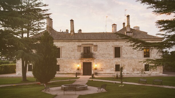 A stone building with a tiled roof, a central entrance, and a well-maintained lawn with benches. - La Posada de Pradorey (Gumiel de Mercado)