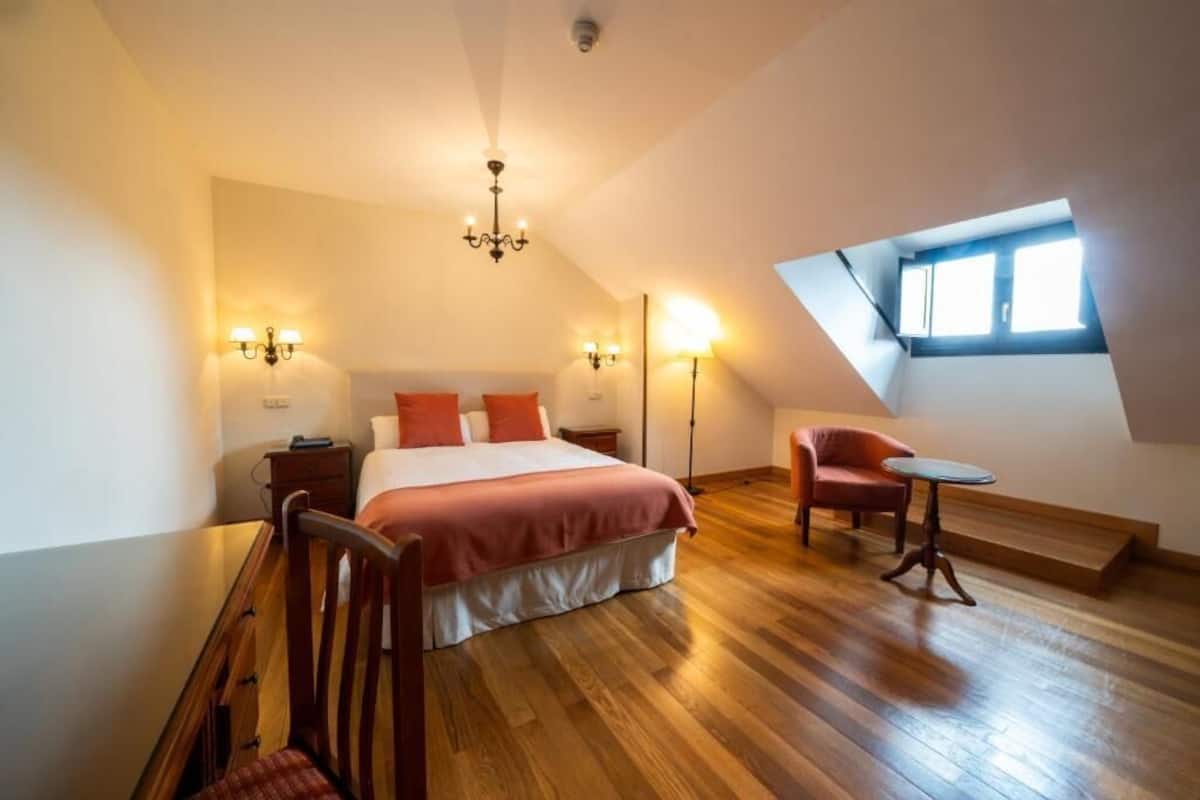 A bedroom with a large bed, a desk, a chair, and a small table. There is a window with a view of the sky.