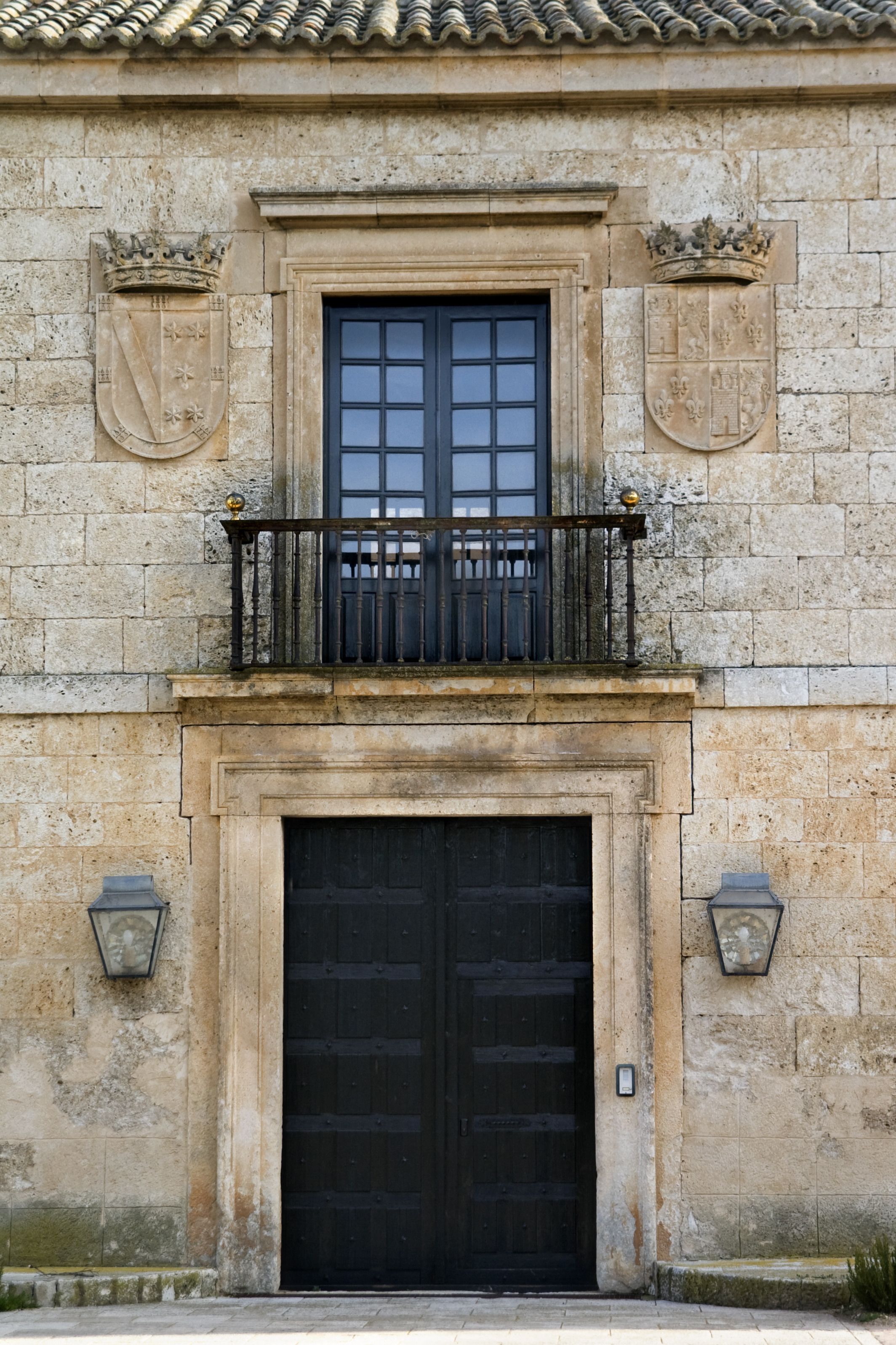 Edifici de pedra amb una porta negra i una finestra, un balcó amb barana i dos escuts al façana.
