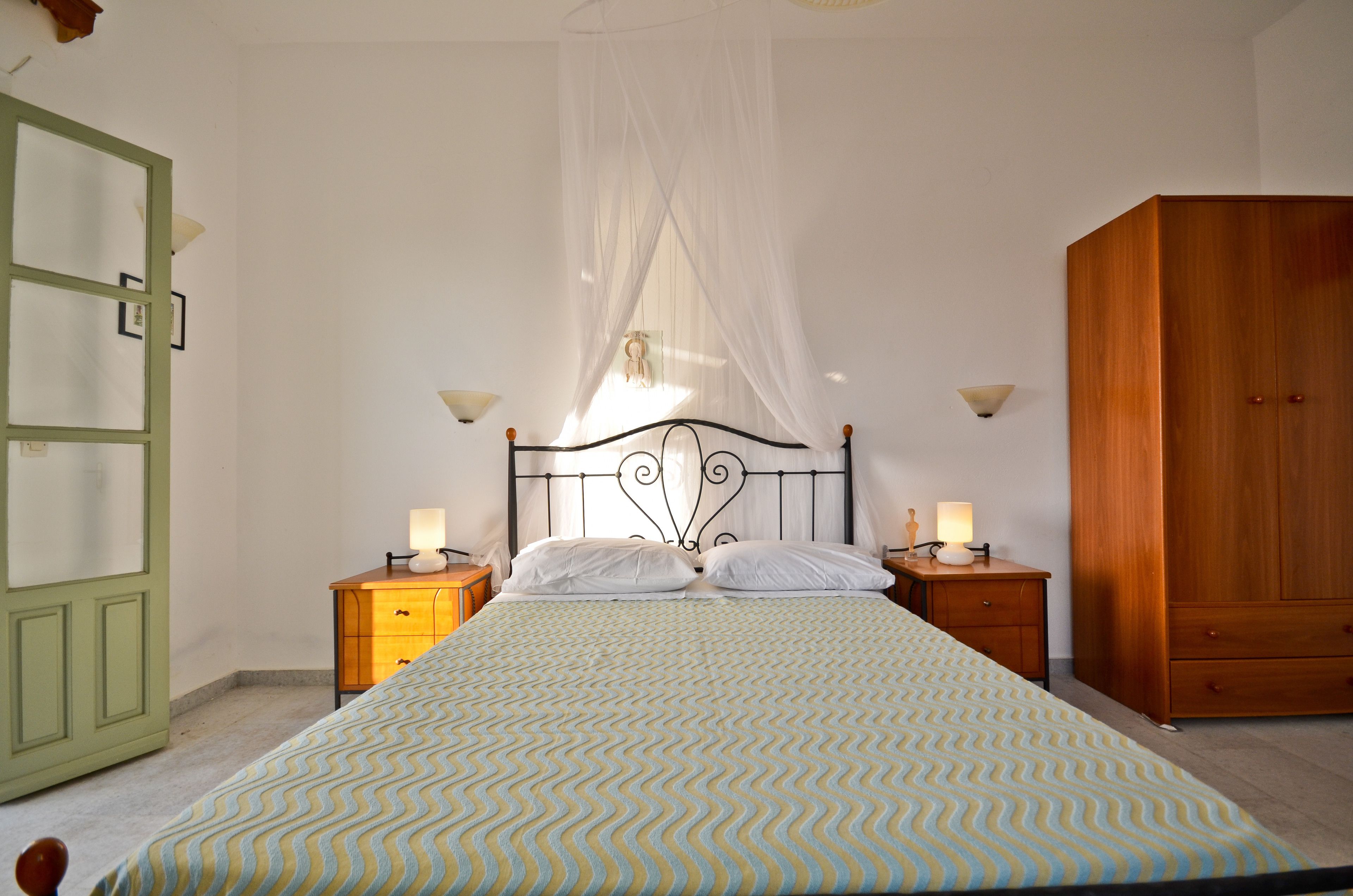 Un dormitorio con cama, mesitas de noche, armario y ventana con cortina translúcida.