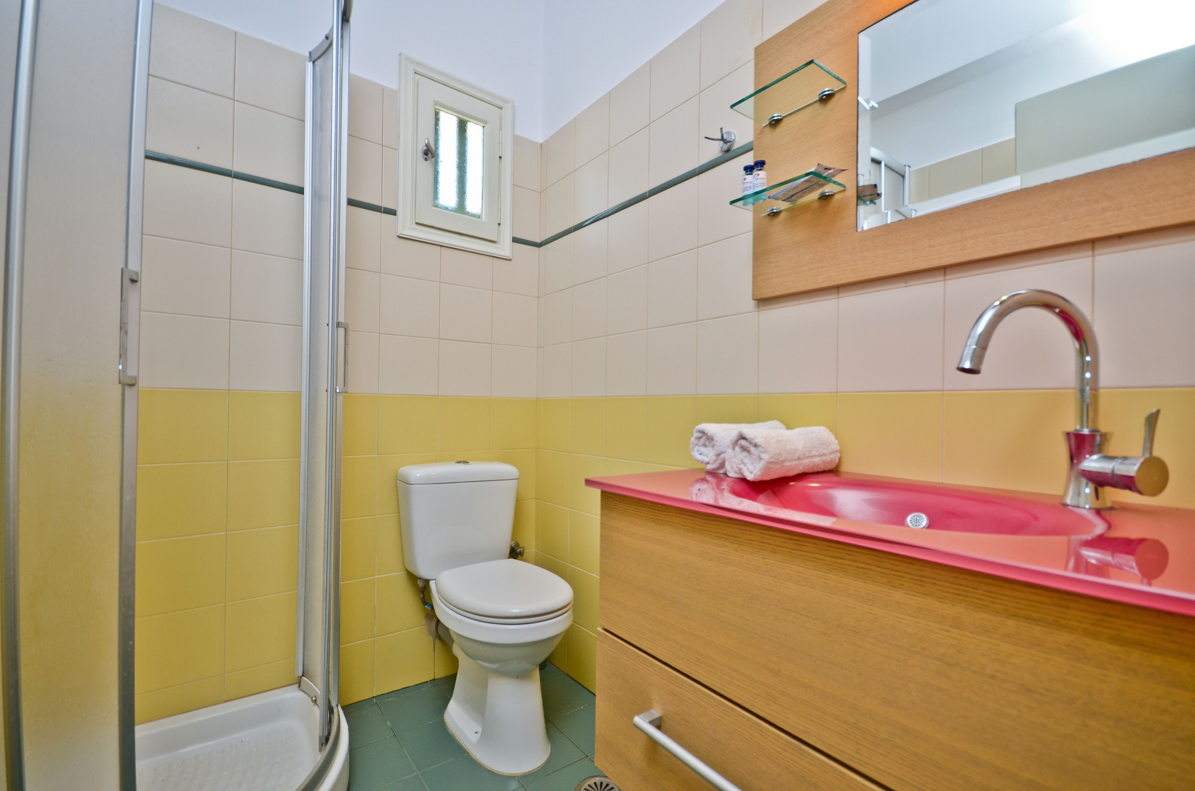 Un baño con pared de azulejos amarillos y blancos, inodoro blanco, mampara de ducha de cristal, tocador de madera con lavabo rojo y un espejo sobre el lavabo.