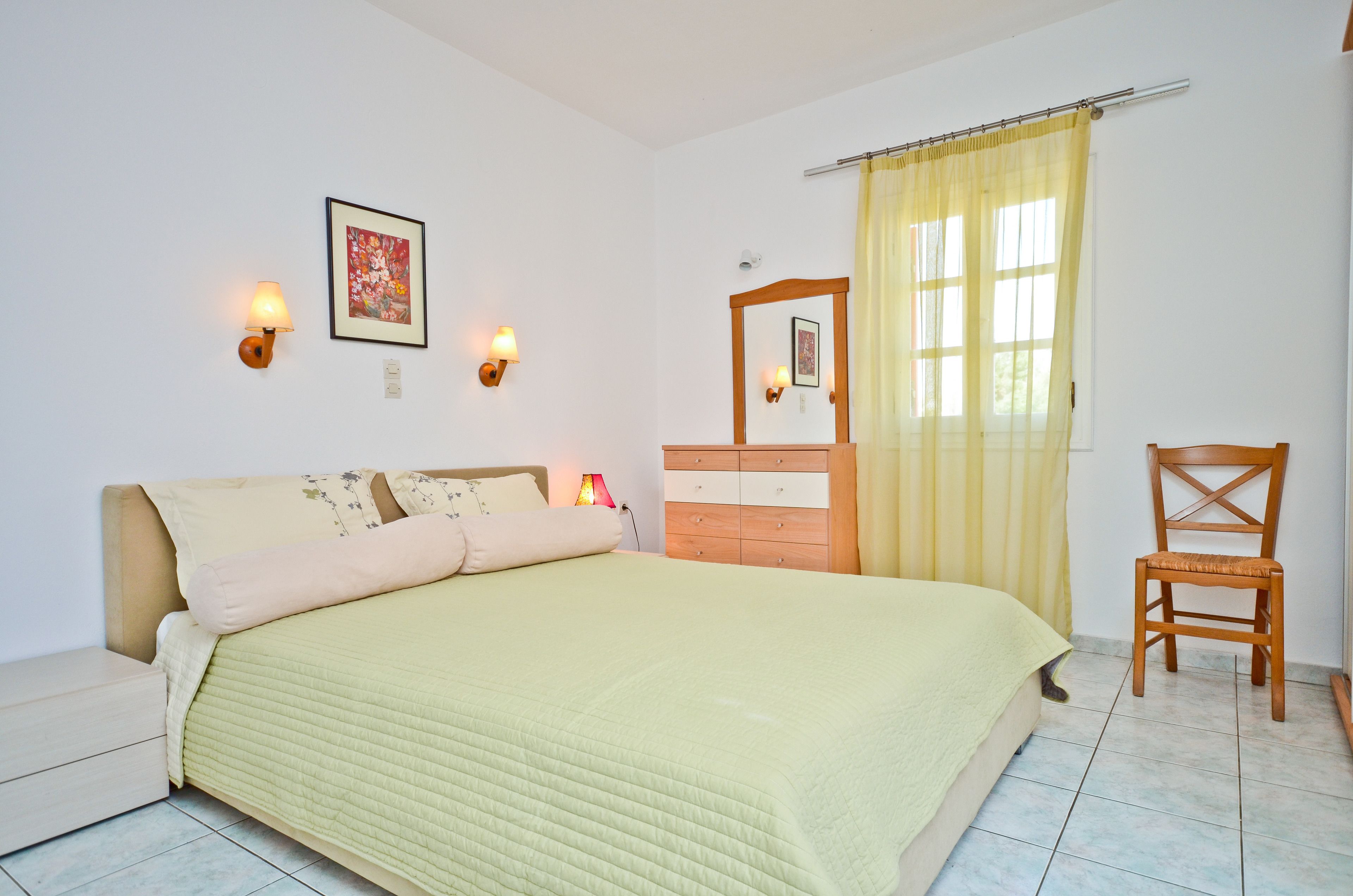Un dormitorio con cama, lámparas de noche, cómoda de madera, una silla y una ventana con cortinas.