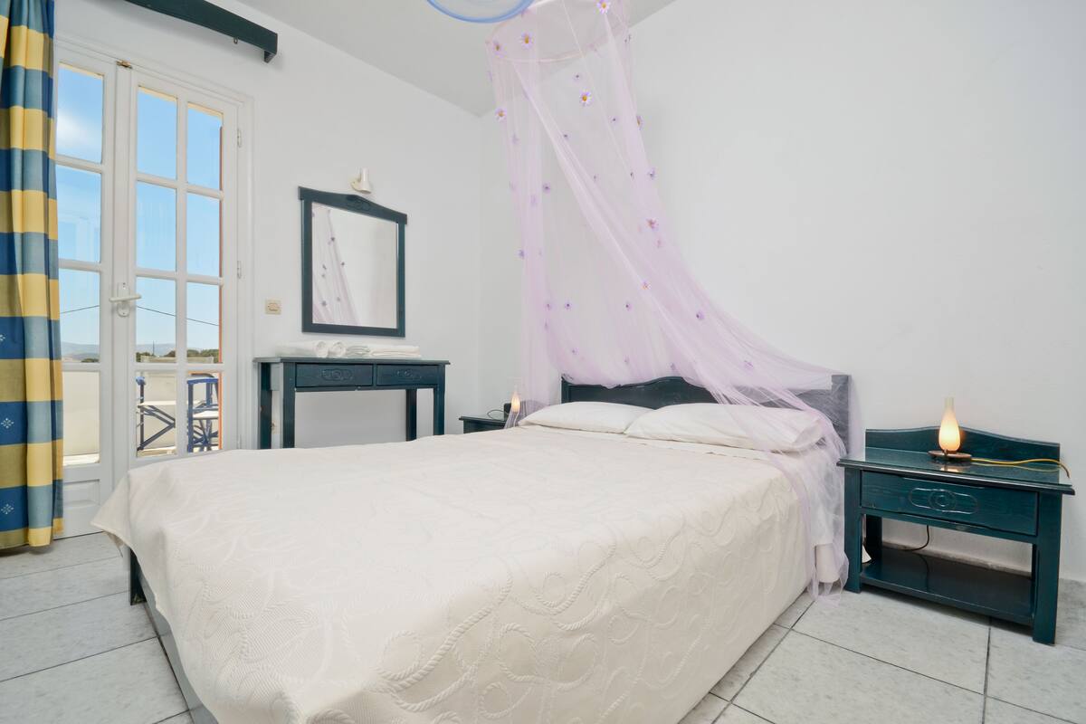 Ein Schlafzimmer mit Bett, Nachttisch, Fenster mit Vorhängen und Blick ins Freie.