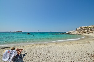 Una playa con agua turquesa cristalina, un promontorio rocoso a la derecha y una persona relajándose en una silla de playa.