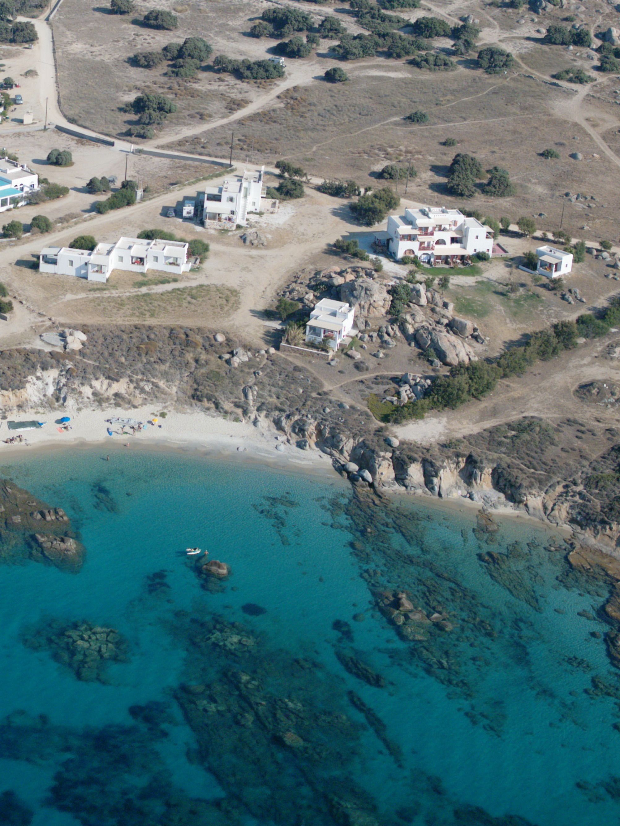 An aerial view of a coastal area with several white buildings, a sandy beach, and rocky terrain.