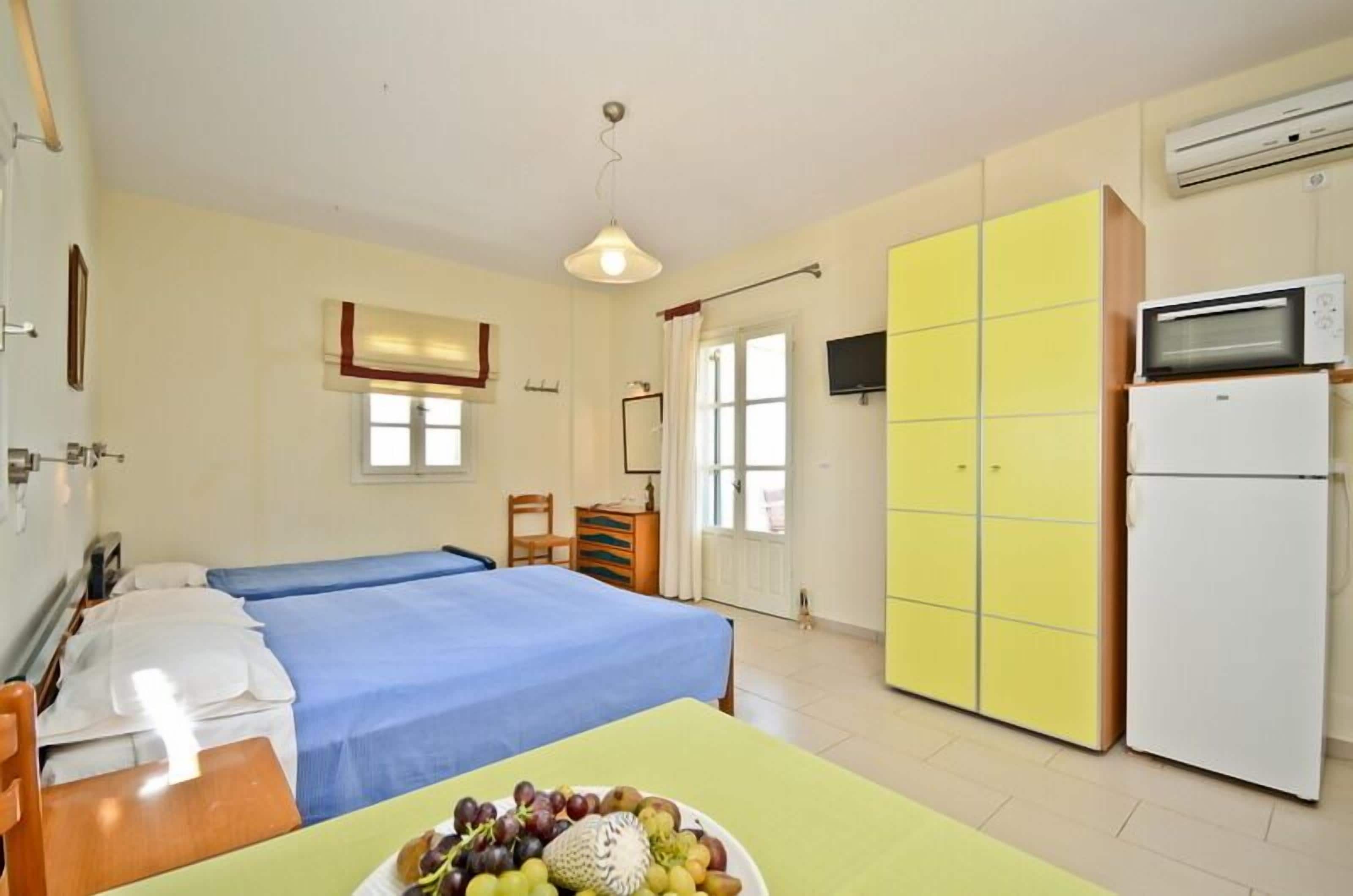 Een hotelkamer met twee bedden, een eettafel met fruit, een gele kast, een witte koelkast en een airconditioner.