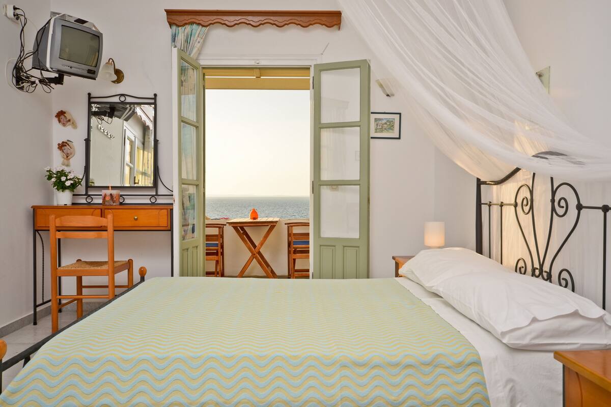 Ein Schlafzimmer mit Bett, Schreibtisch, Stuhl, Fernseher und Blick auf das Meer durch eine offene Tür.