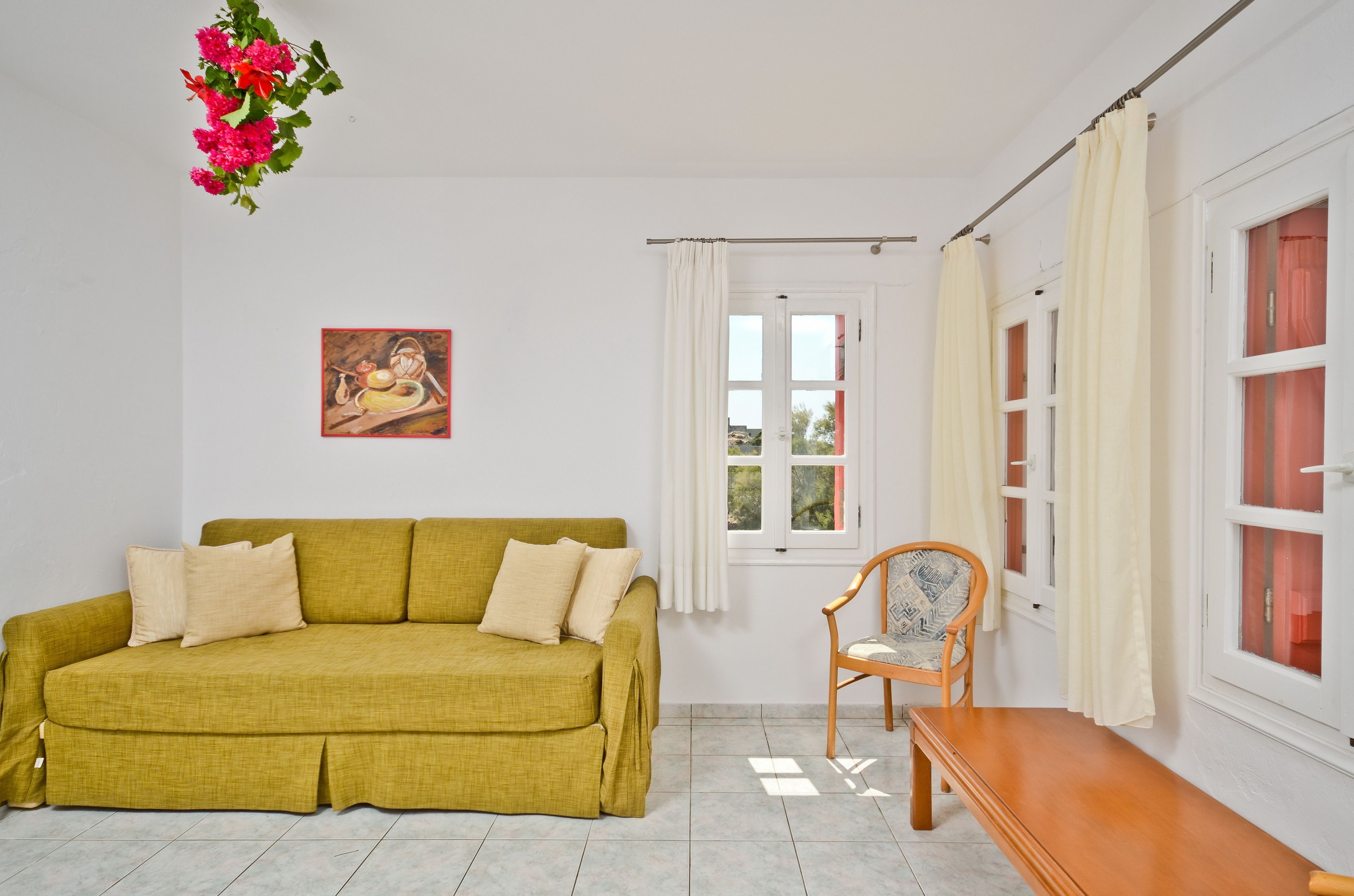 Una habitación con un sofá amarillo, un banco de madera, una silla, una ventana con cortinas y un cuadro en la pared.