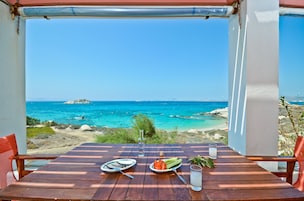 Una mesa de madera con platos de comida y un vaso de agua, con vista a una playa de aguas azules y terreno rocoso.