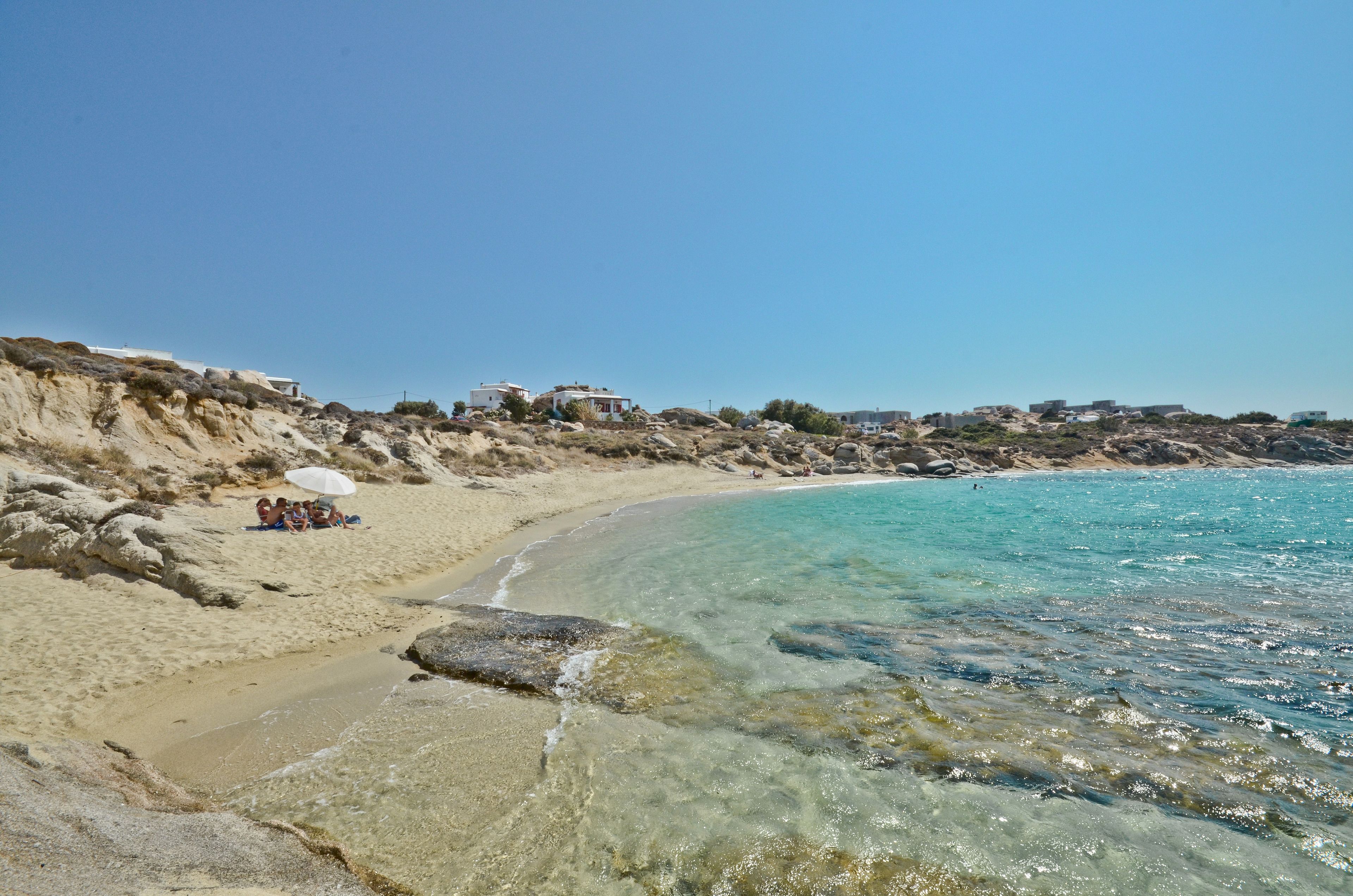 A beach with clear turquoise water, rocky shoreline, and buildings on the cliff.